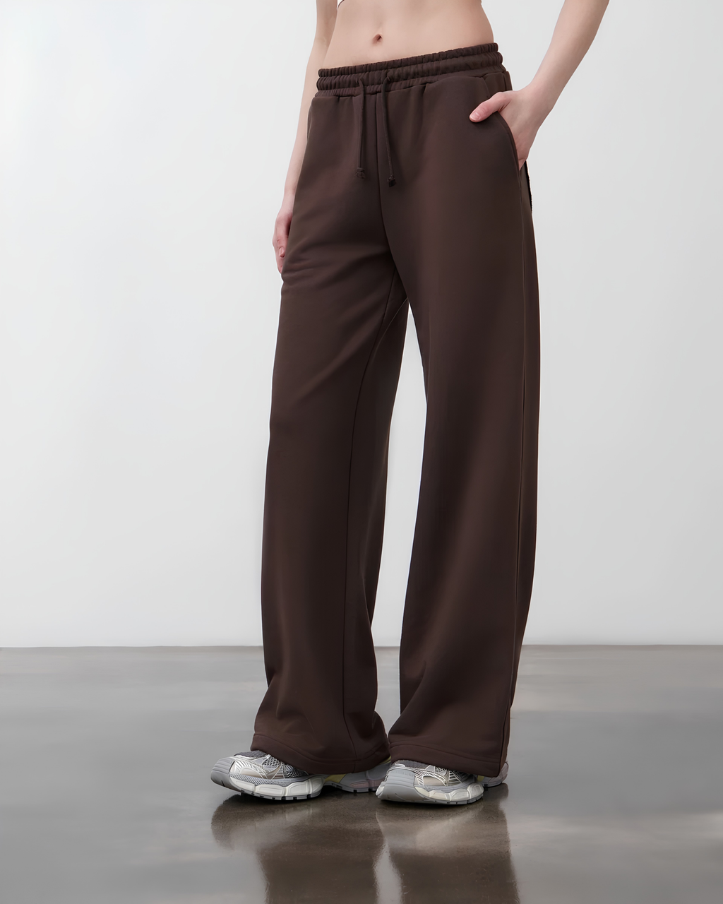 A person wearing loose, wide-leg brown drawstring sweatpants with side pockets, paired with gray sneakers, standing in a studio setting.