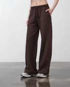 A person wearing loose, wide-leg brown drawstring sweatpants with side pockets, paired with gray sneakers, standing in a studio setting.