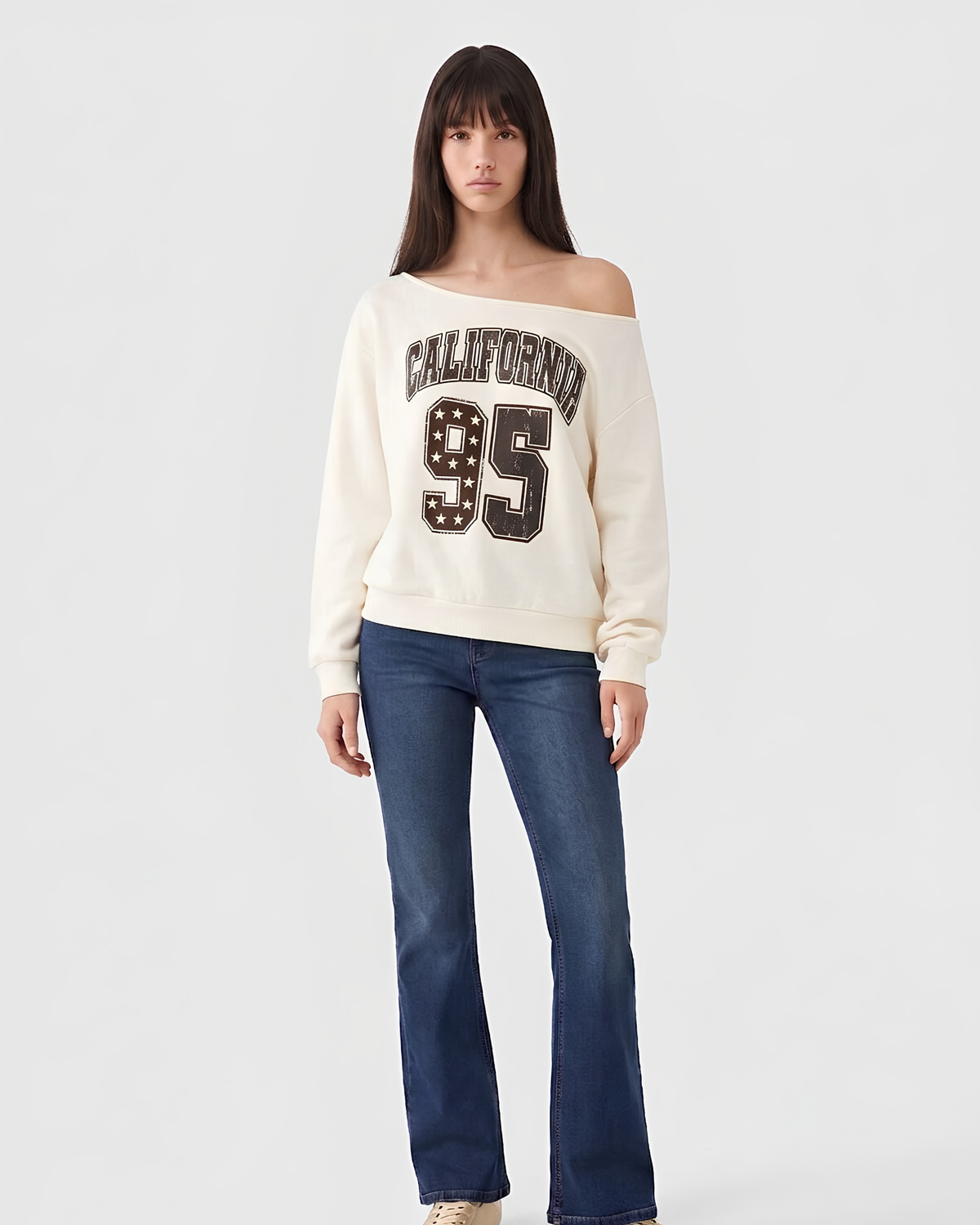 A woman modeling an off-the-shoulder cream sweatshirt with a 'CALIFORNIA 95' graphic on the front, paired with dark blue jeans, against a light gray studio background.