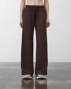 A model from the waist down wearing dark brown wide-leg sweatpants with a drawstring waistband, paired with silver sneakers, against a neutral studio background.