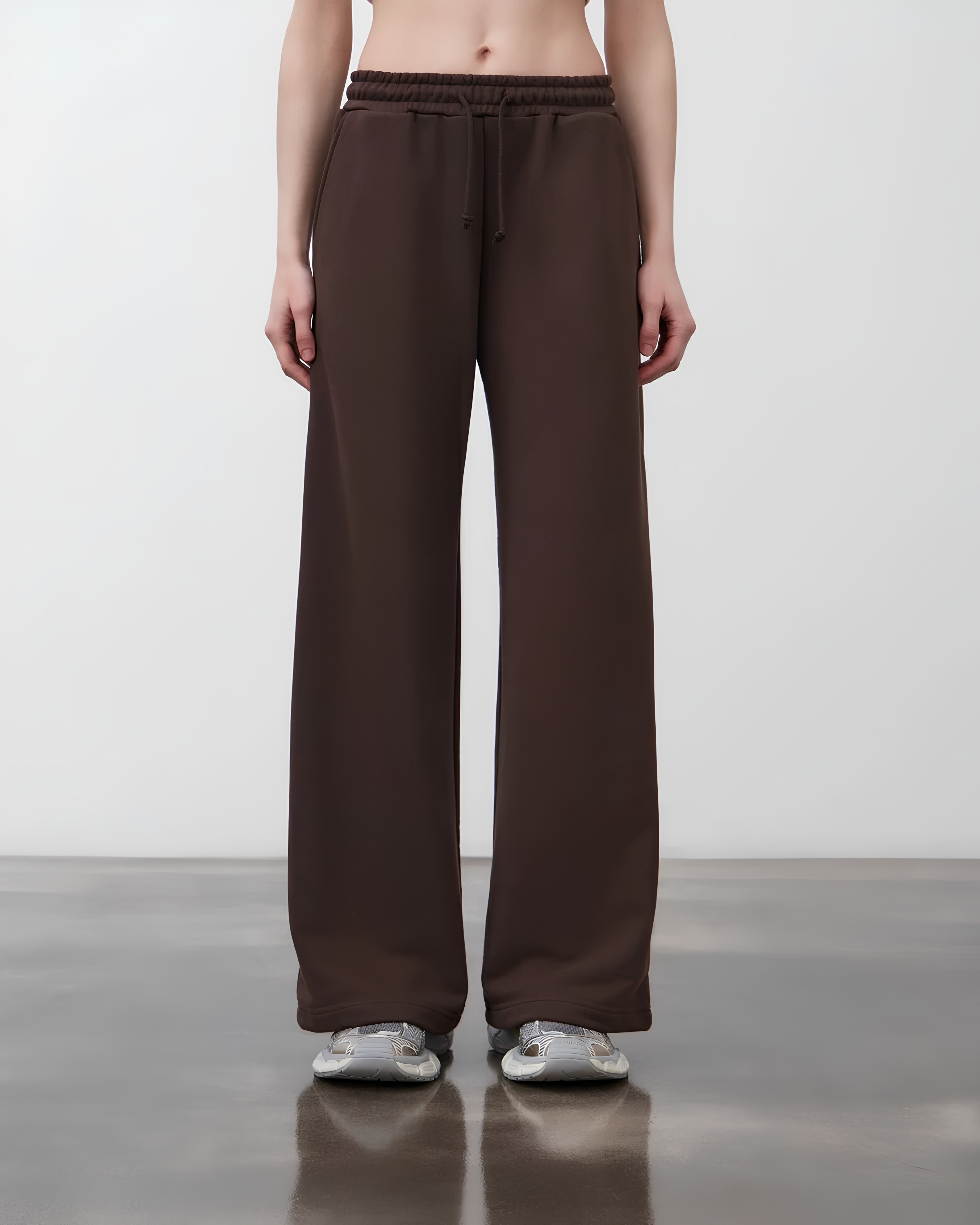 A model from the waist down wearing dark brown wide-leg sweatpants with a drawstring waistband, paired with silver sneakers, against a neutral studio background.