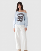A model wearing a light blue off-the-shoulder sweatshirt with 'CALIFORNIA 95' printed on the front, paired with white wide-leg pants, against a plain light gray background.