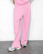 Pink wide-leg sweatpants worn by a model, paired with silver sneakers, shown from the waist down against a white wall and dark floor.