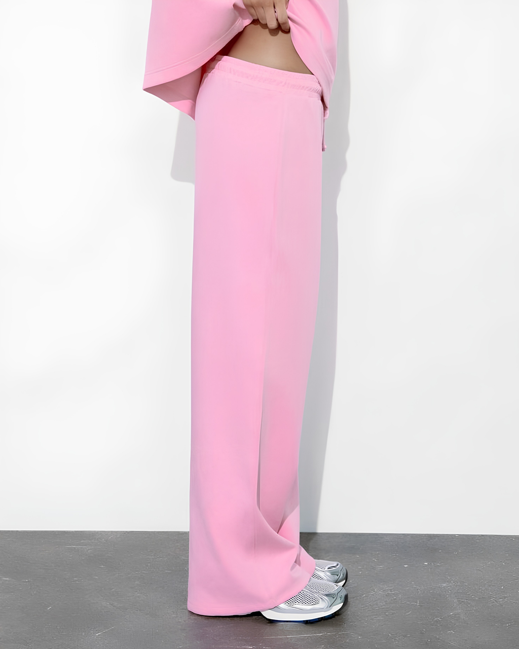 A person wearing bright pink wide-leg pants and a matching pink top, standing against a plain white wall with a dark gray floor. The person is shown from the waist down, wearing silver sneakers.
