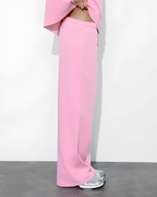 A person wearing bright pink wide-leg pants and a matching pink top, standing against a plain white wall with a dark gray floor. The person is shown from the waist down, wearing silver sneakers.