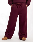 A person wearing loose, wide-leg sweatpants in a deep burgundy or plum color, paired with a matching cropped sweatshirt and tan shoes, shown against a plain white studio background.