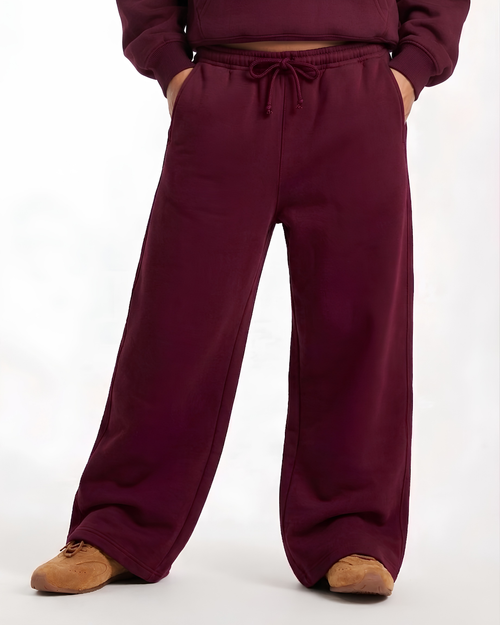 A person wearing loose, wide-leg sweatpants in a deep burgundy or plum color, paired with a matching cropped sweatshirt and tan shoes, shown against a plain white studio background.