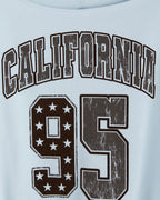 Close-up of a light blue T-shirt with a distressed dark brown graphic reading 'CALIFORNIA 95' on the chest.