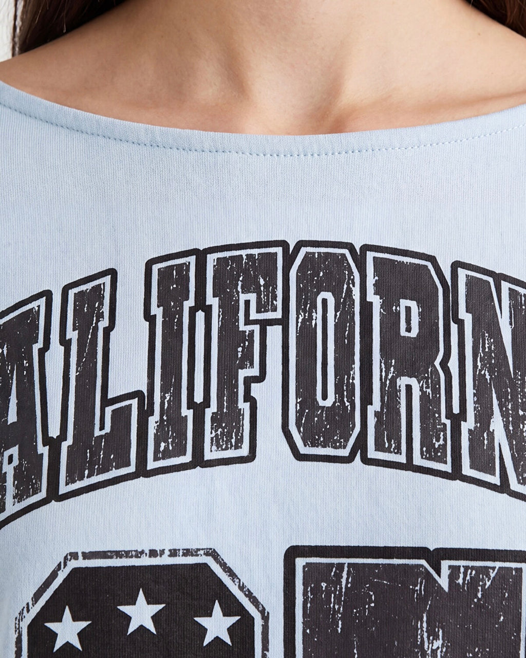 Close-up of a person wearing a light blue off-the-shoulder graphic top with distressed black and white collegiate-style lettering.