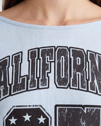 Close-up of a person wearing a light blue off-the-shoulder graphic top with distressed black and white collegiate-style lettering.