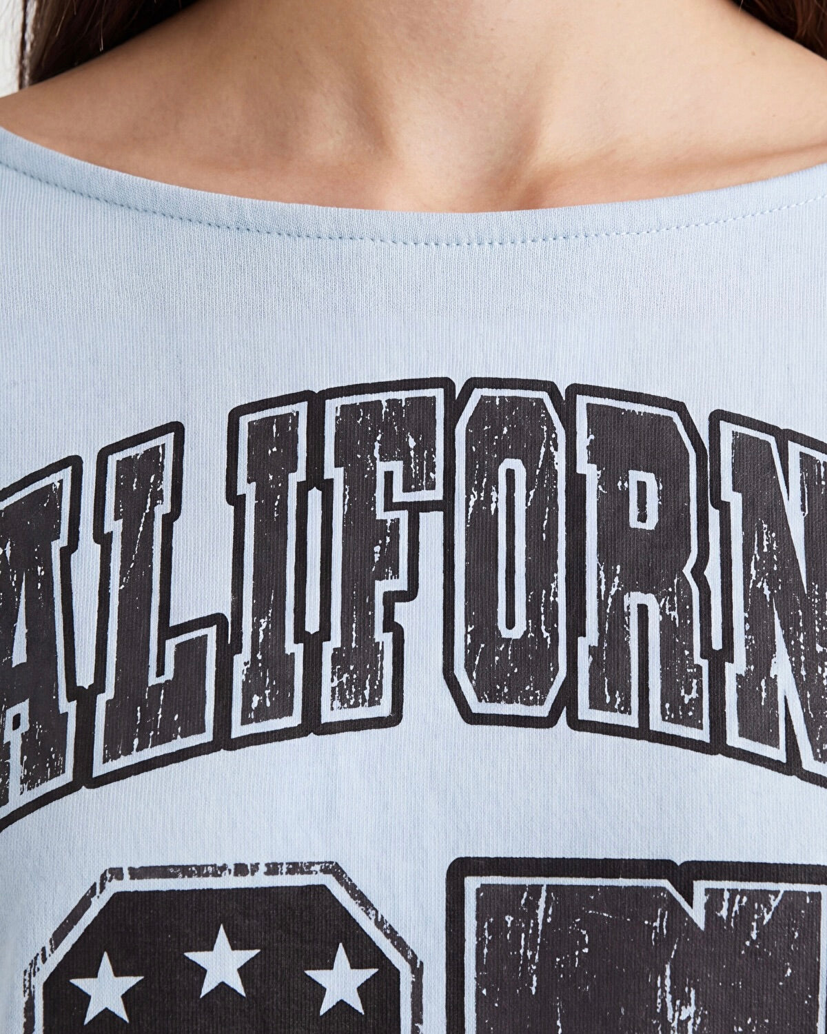 Close-up of a person wearing a light blue off-the-shoulder graphic top with distressed black and white collegiate-style lettering.