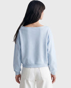A woman shown from the back wearing a light blue long-sleeve cropped sweatshirt and white pants against a plain light gray studio background.
