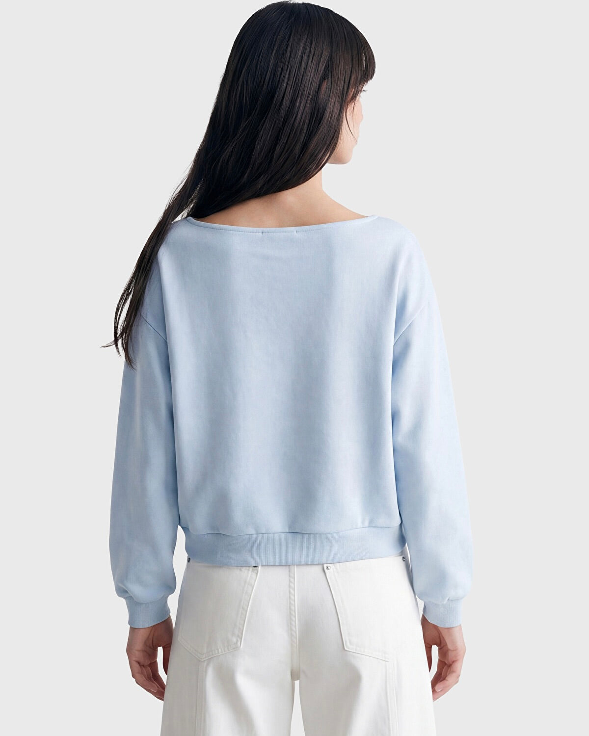 A woman shown from the back wearing a light blue long-sleeve cropped sweatshirt and white pants against a plain light gray studio background.