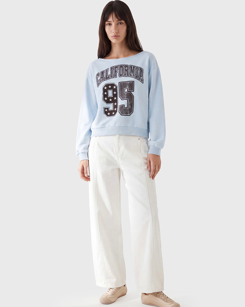 A woman modeling a light blue crewneck sweatshirt with 'CALIFORNIA 95' printed on the front, paired with white wide-leg pants and beige sneakers, against a light gray studio background.
