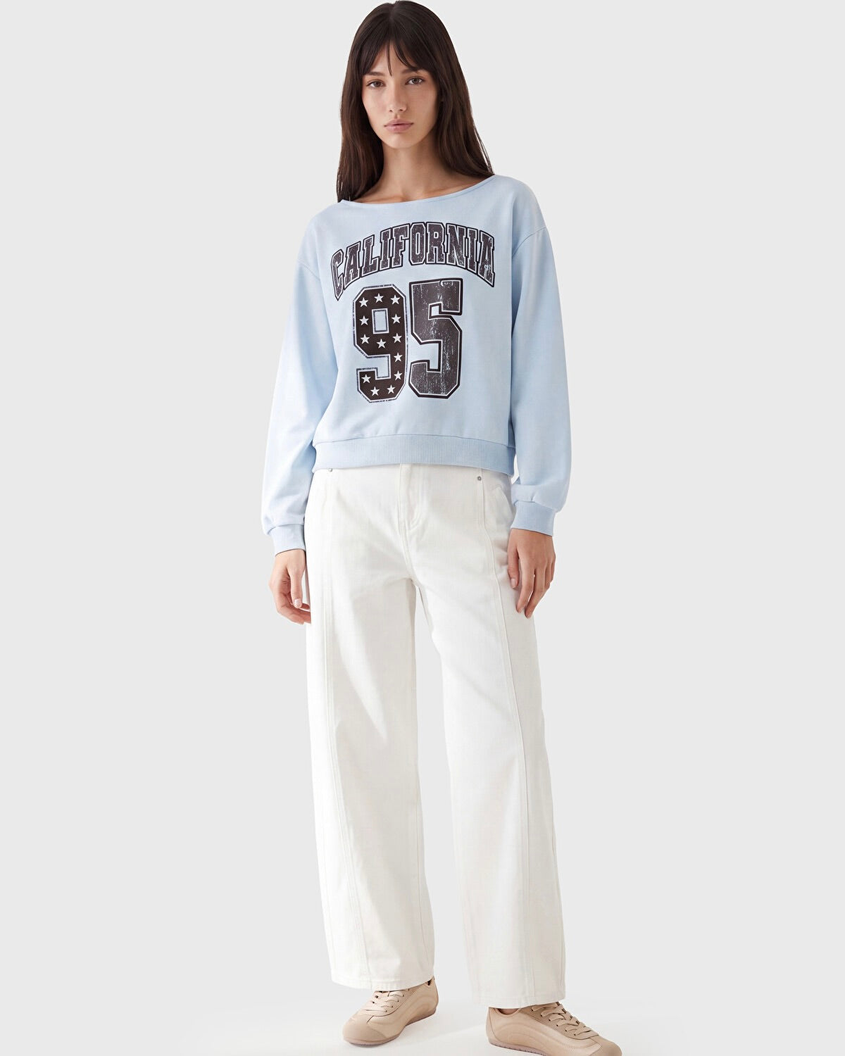 A woman modeling a light blue crewneck sweatshirt with 'CALIFORNIA 95' printed on the front, paired with white wide-leg pants and beige sneakers, against a light gray studio background.