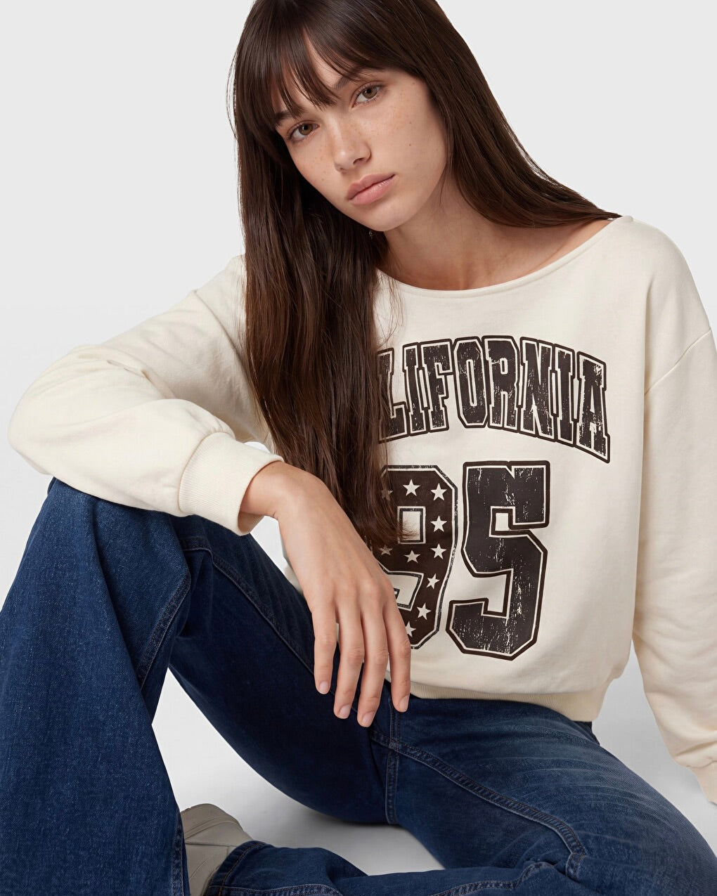A woman wearing a cream-colored crewneck sweatshirt with a distressed black 'CALIFORNIA 95' graphic, paired with dark blue jeans, posed against a light gray studio background.