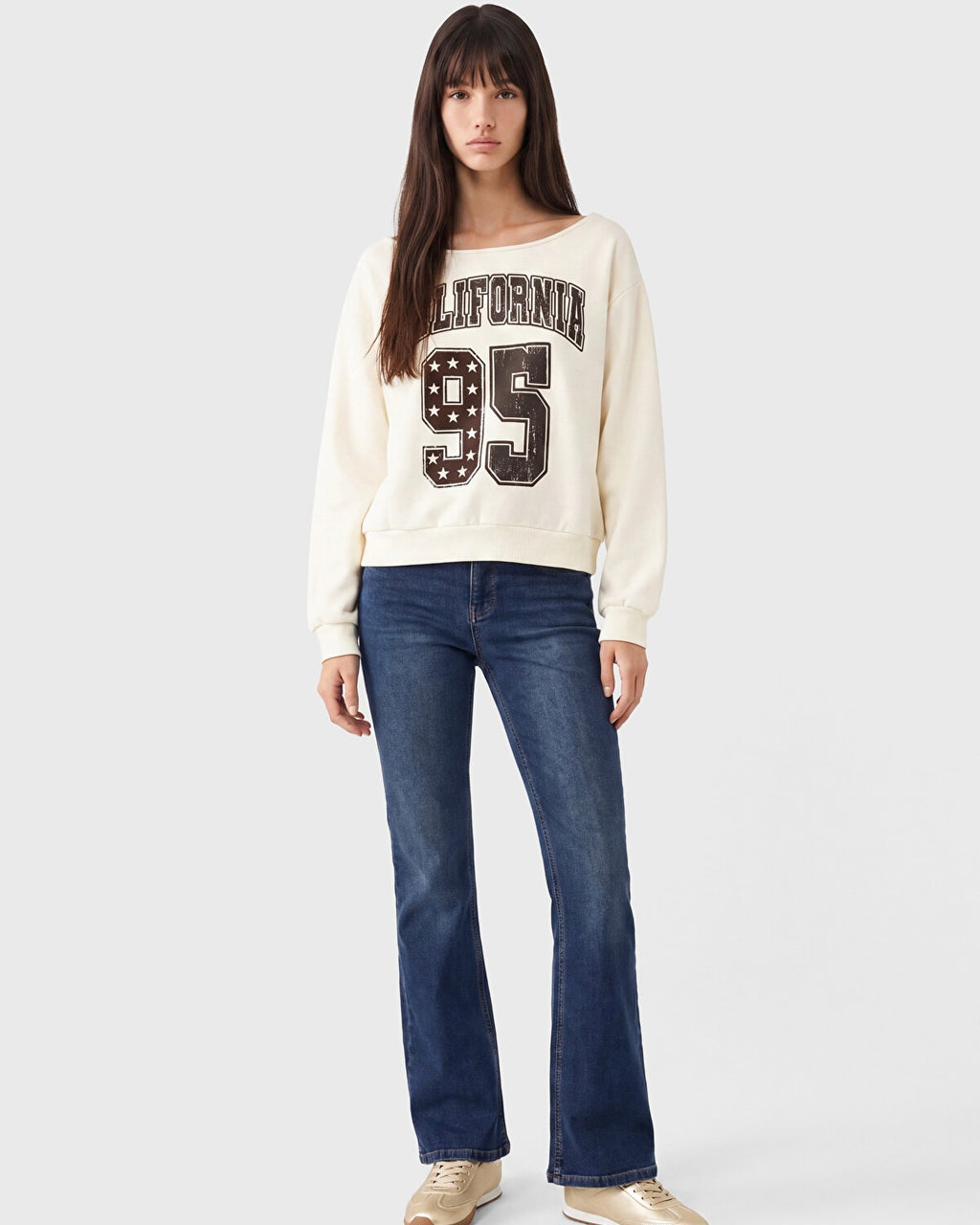 A woman modeling a cream-colored crewneck sweatshirt with 'CALIFORNIA 95' printed on the front in black and white, paired with dark blue jeans and beige sneakers, against a light gray studio background.