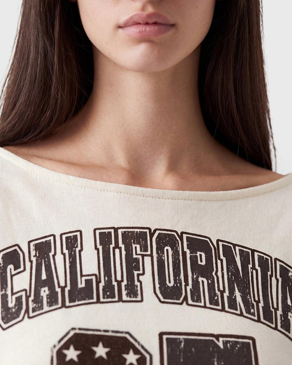 Close-up of a model wearing a cream-colored off-the-shoulder t-shirt with a distressed dark brown graphic that reads 'CALIFORNIA'.