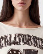 Close-up of a model wearing a cream-colored off-the-shoulder t-shirt with a distressed dark brown graphic that reads 'CALIFORNIA'.