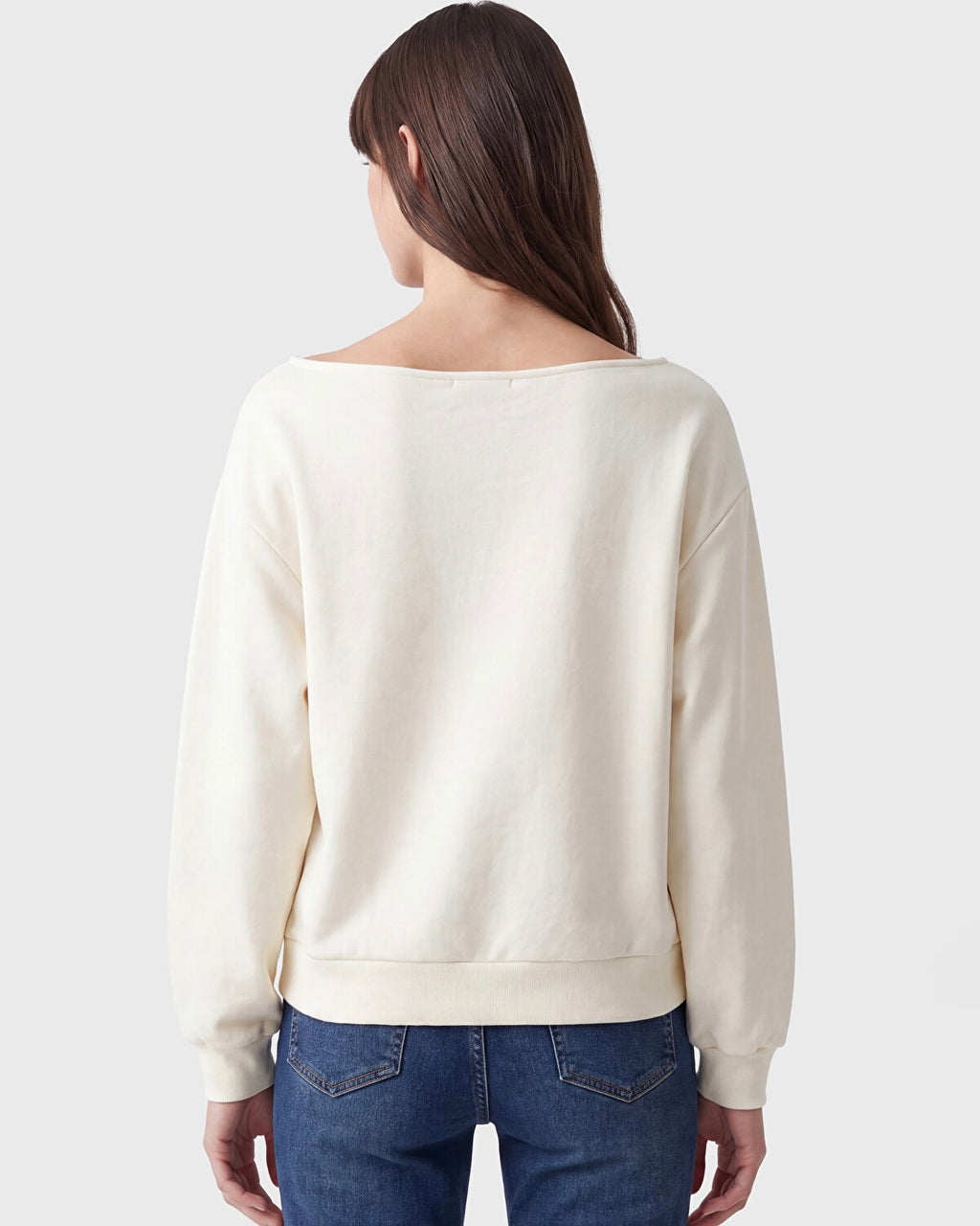 A woman viewed from the back wearing a cream-colored wide-neck sweatshirt and blue jeans against a light gray studio background.