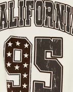 Close-up of a distressed varsity-style graphic print featuring the word 'CALIFORNIA' in arched block letters and the number '95' with white stars on a dark brown and off-white background.