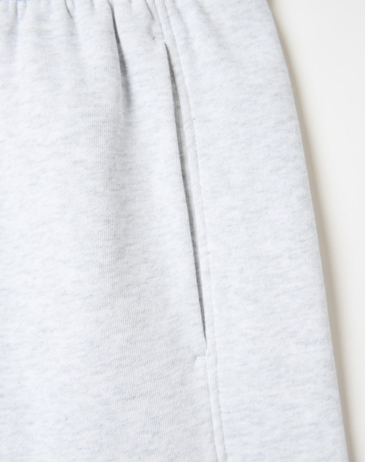 Close-up of light heather gray fabric, likely sweatpants or a soft garment, showing a vertical seam and a side pocket on a plain light background.