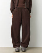 A person wearing dark brown wide-leg sweatpants with an elastic drawstring waist, paired with a matching brown top and gray sneakers, standing against a plain light gray wall.