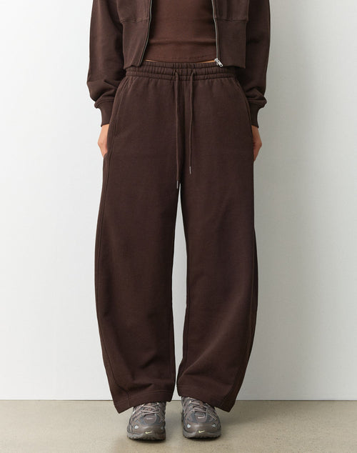 A person wearing dark brown wide-leg sweatpants with an elastic drawstring waist, paired with a matching brown top and gray sneakers, standing against a plain light gray wall.