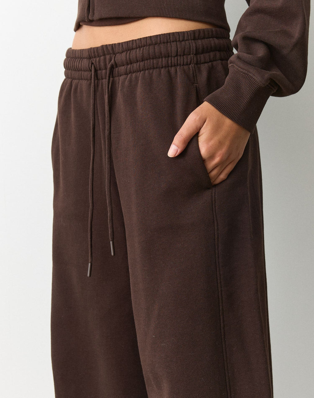 Close-up of a person wearing dark brown wide-leg sweatpants with an elastic drawstring waist and side pockets, paired with a matching cropped top against a light gray studio background.