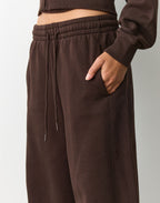 Close-up of a person wearing dark brown wide-leg sweatpants with an elastic drawstring waist and side pockets, paired with a matching cropped top against a light gray studio background.